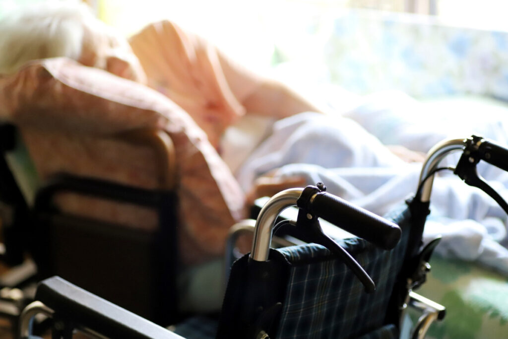 高齢女性がベッドで横になっており、手前に車椅子が写っている写真